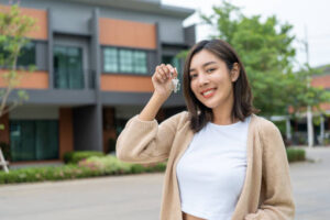 Woman buying or rent new home she holding key front of new house. Surprise happy young asian woman giving house key and smile to rent or purchase apartment home. Moving relocation concept.